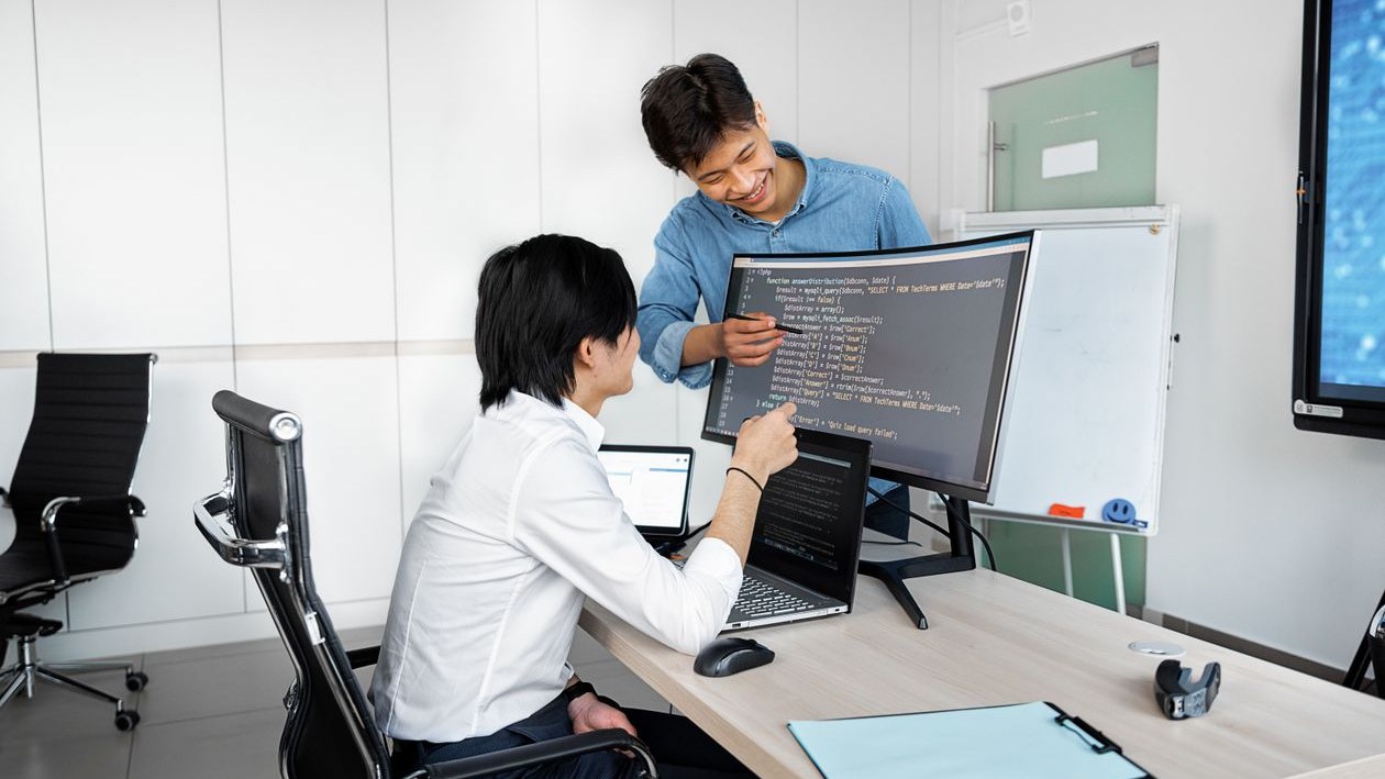 A young man at an office desk works on code at a large monitor while a smiling colleague stands beside him and points at the screen.